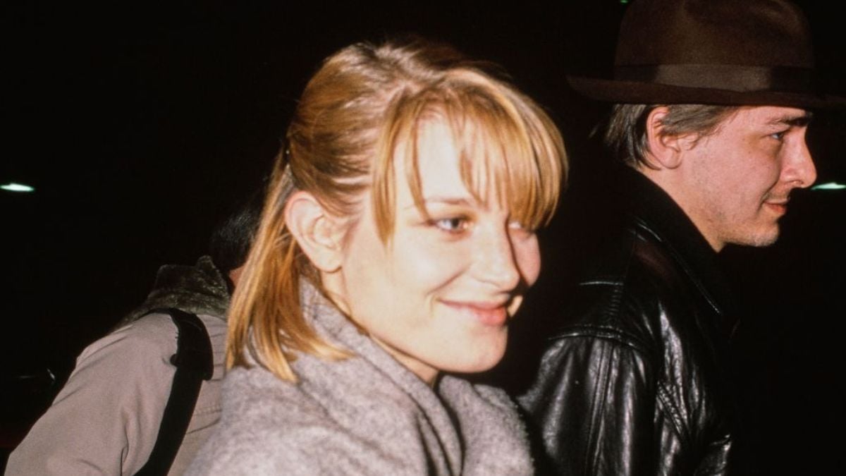 A woman with blonde hair and bangs smiles while wearing a gray coat. A man in a brown hat and black leather jacket stands beside her. The background is dark and out of focus.
