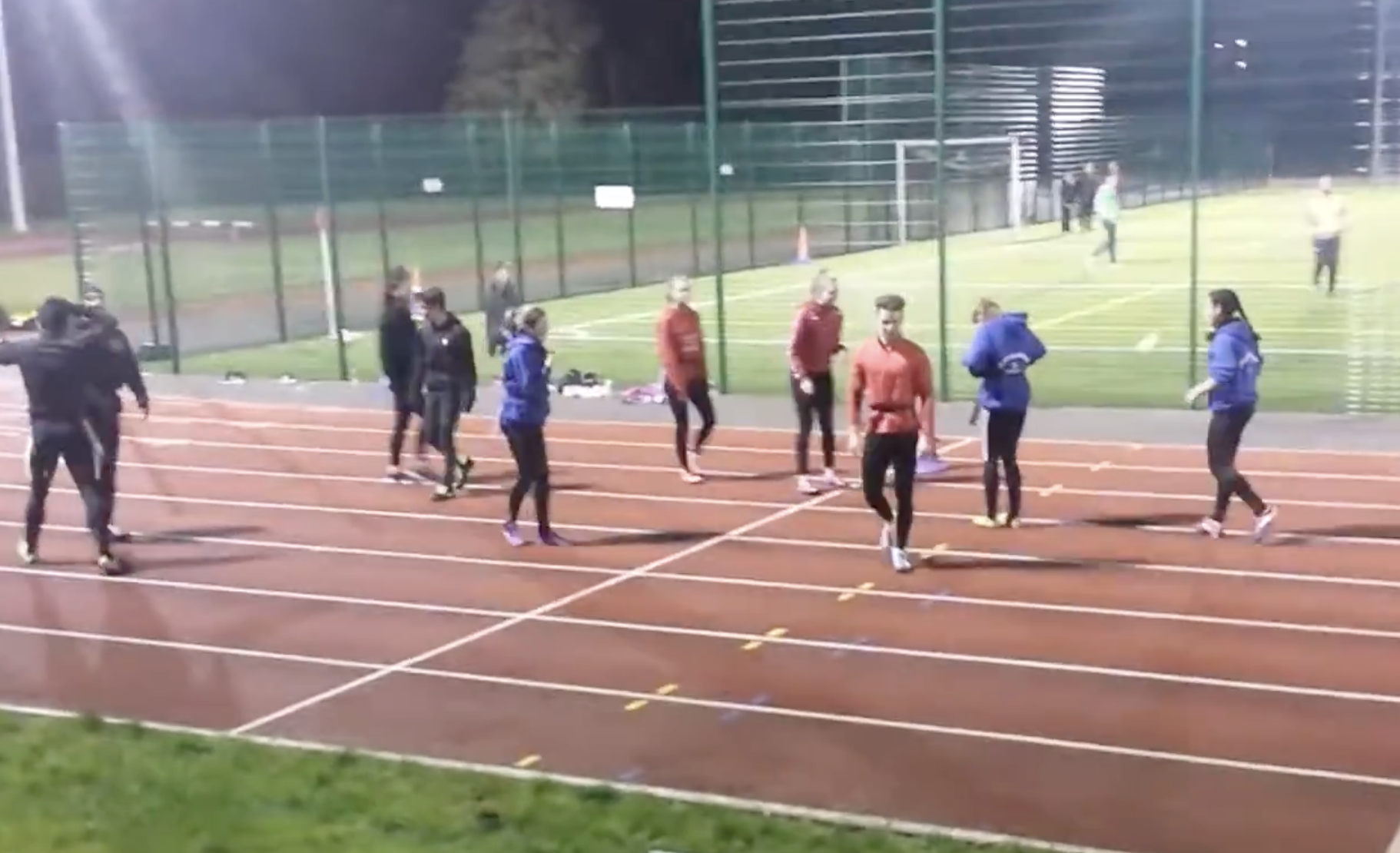A group of athletes in colorful tracksuits walk across a running track at night.