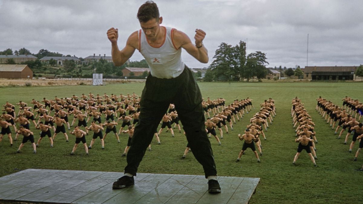 A man leads a large group of people in exercise on a grassy field, standing on a platform with arms bent, while rows of participants mirror his movements behind him.
