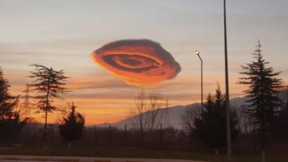 A dramatic, orange lenticular cloud shaped like a UFO hovers over treetops at sunset, with mountains silhouetted in the background.