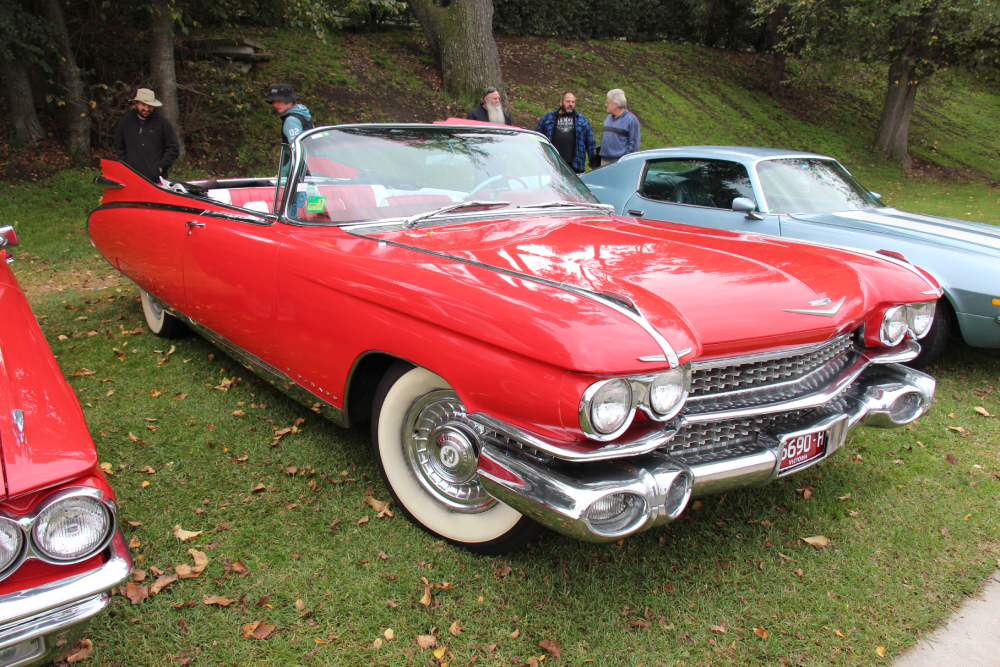 A red vintage Cadillac convertible parked on grass next to a light blue coupe, with several people standing in the background.