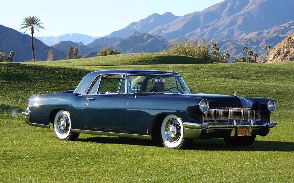 A vintage dark blue Continental Mark II coupe parked on a golf course with mountains in the background.