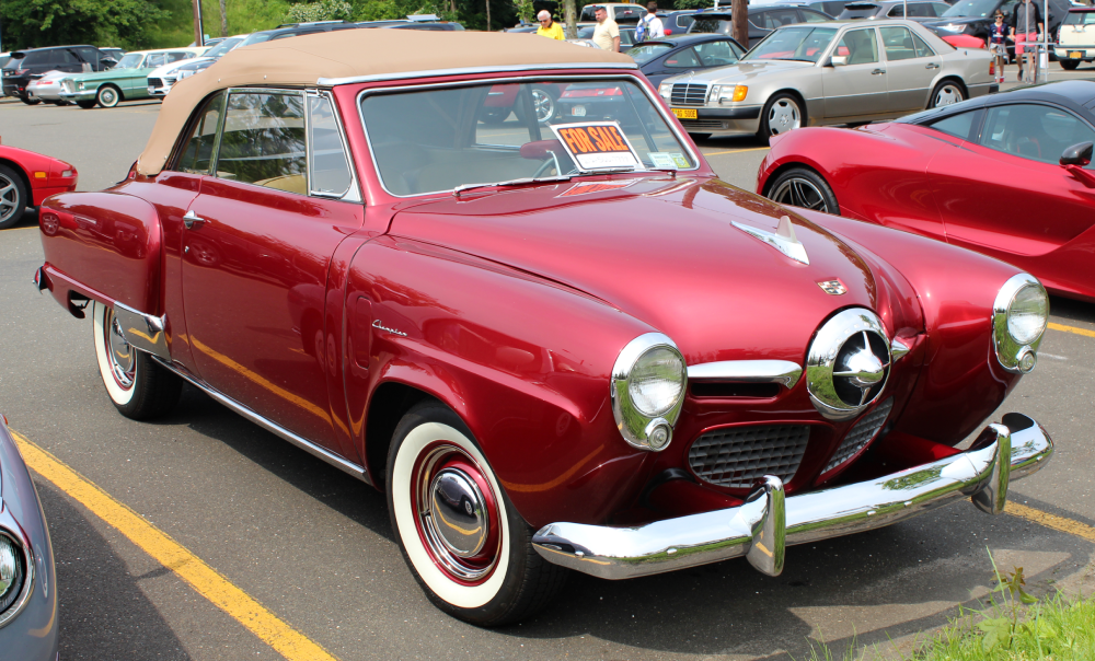 A vintage red Studebaker Champion convertible with a tan top parked in a lot, featuring a “For Sale” sign in the windshield.