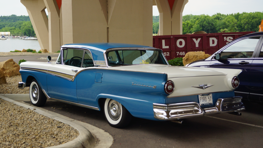 A blue and white 1957 Ford Fairlane 500 parked in a lot near a waterfront.