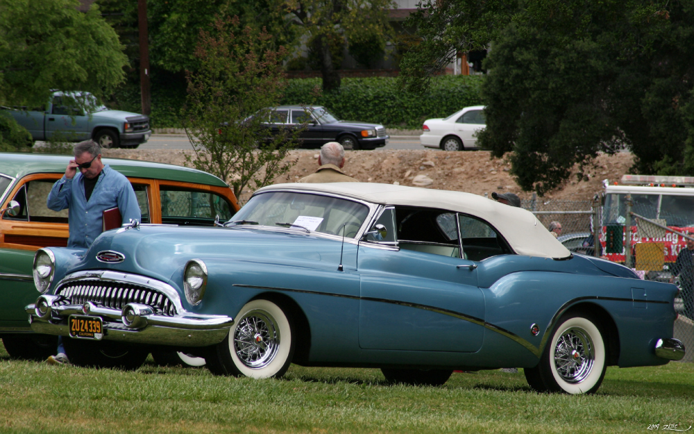 A light blue vintage Buick convertible with a tan roof parked on grass, with a man in a blue shirt standing beside it.