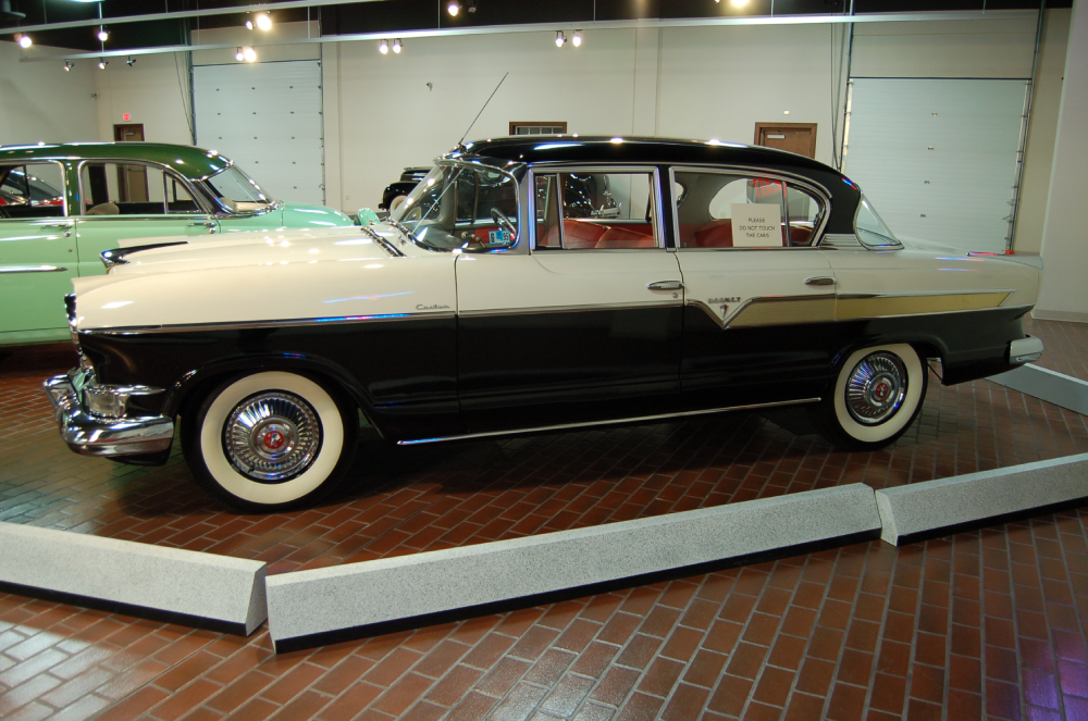 A black and white vintage sedan parked on a brick floor in a showroom, with a partial view of a green vintage car behind it.