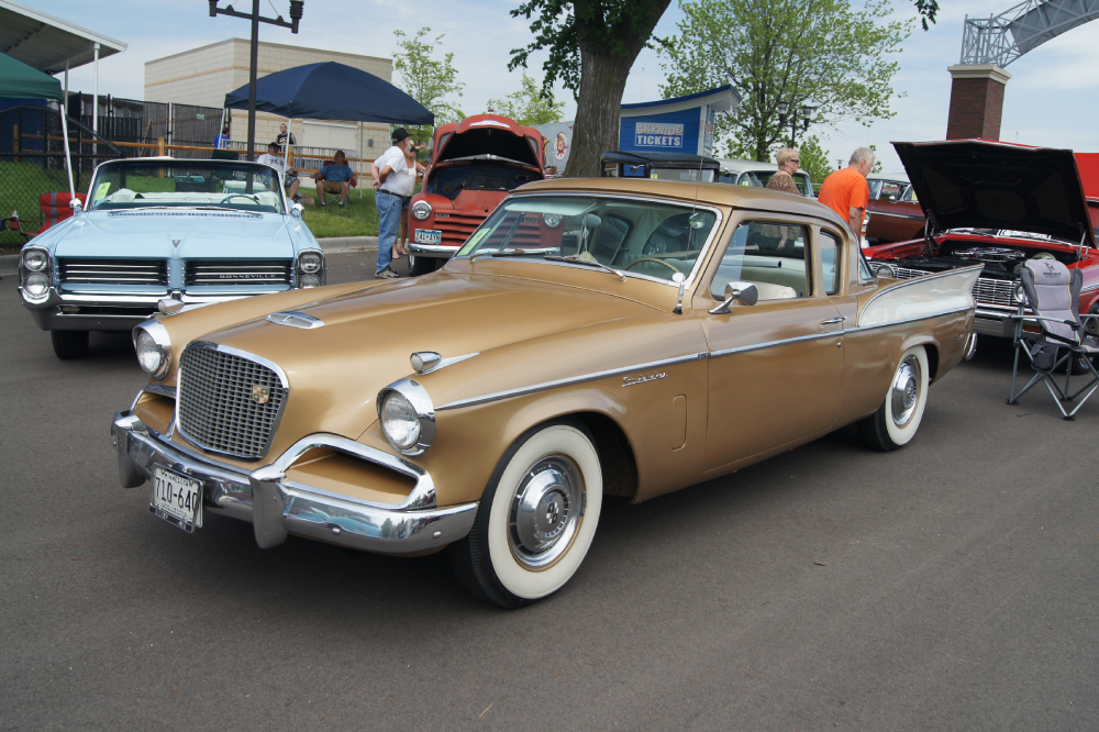 A gold vintage Studebaker coupe with whitewall tires parked at a car show, with other classic cars visible in the background.
