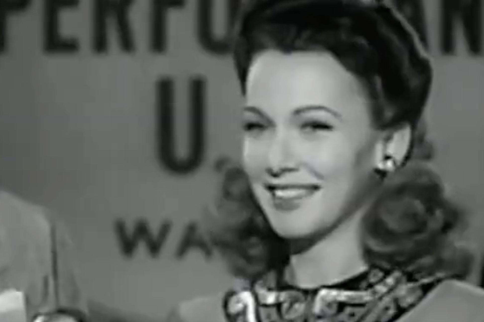 A smiling woman with curled hair wearing a decorative collar, in front of a sign that reads “PERFORMING U.S.”