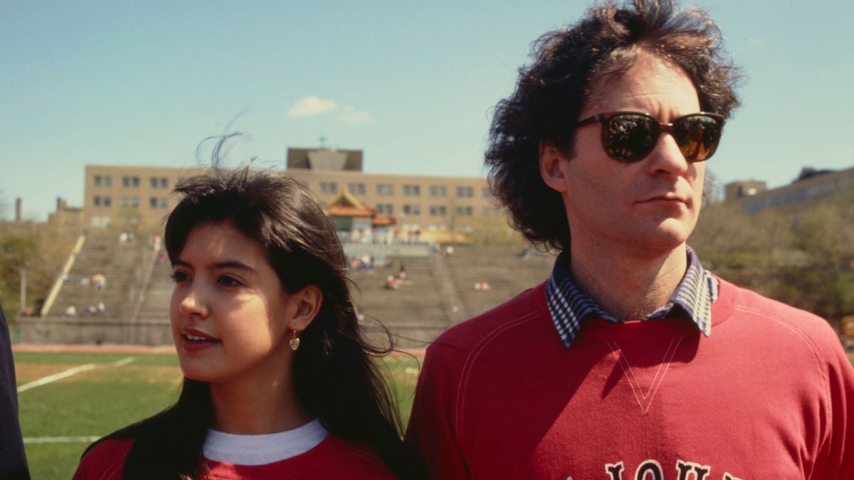 A young woman and a man wearing sunglasses, both in red sweaters, stand outdoors on a sunny day with empty bleachers and a building in the background.