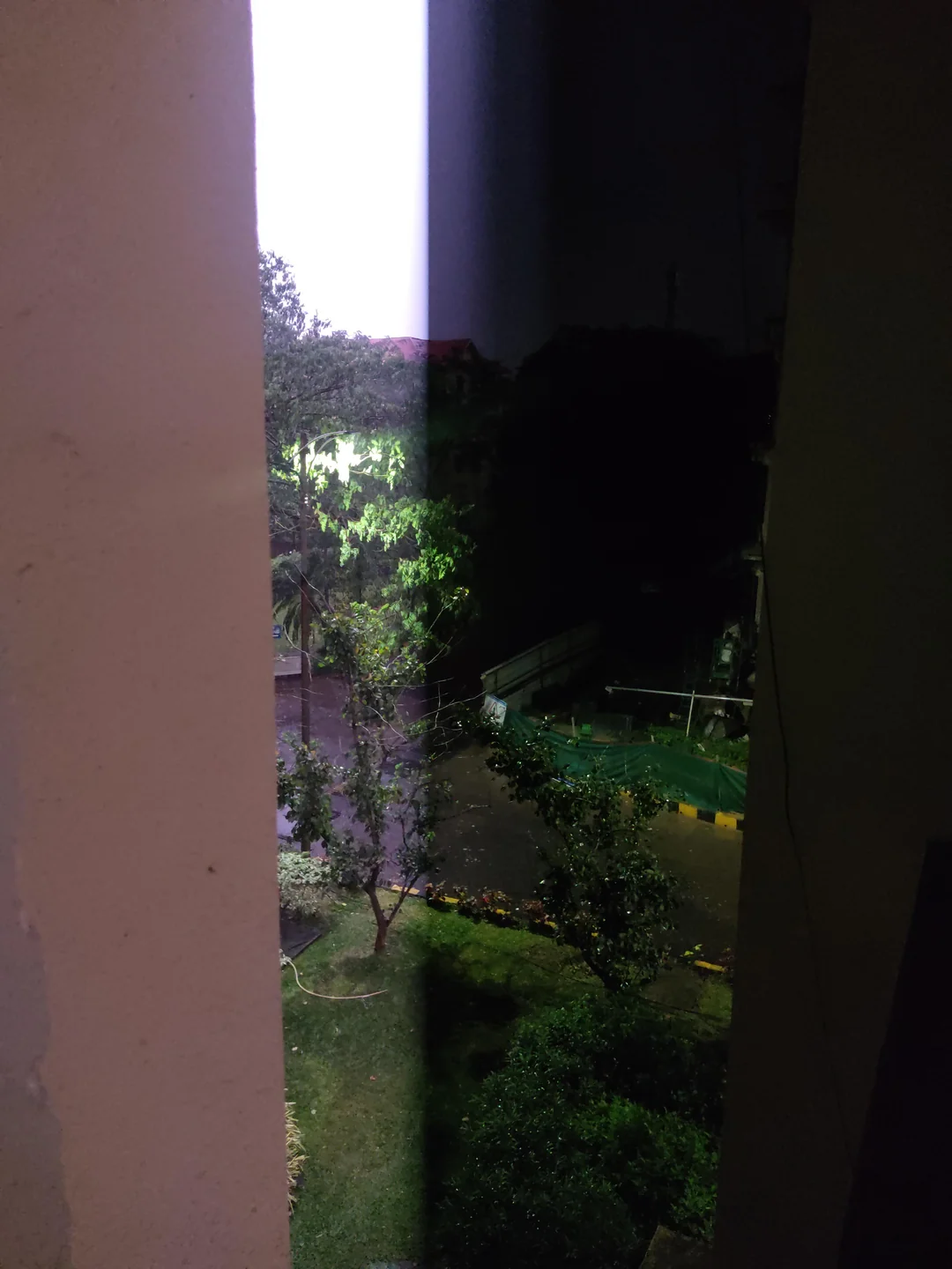 A dark outdoor scene at night viewed through a narrow opening between two walls, showing trees and a patch of grass illuminated by a light.