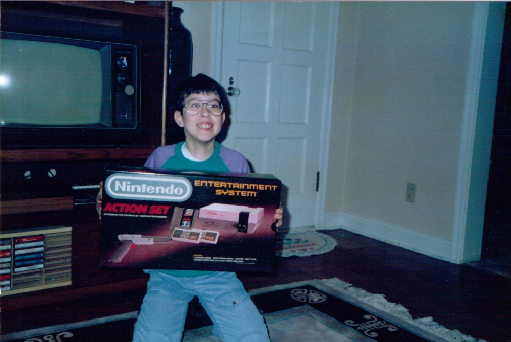 A young boy wearing glasses excitedly holds a boxed Nintendo Entertainment System Action Set.