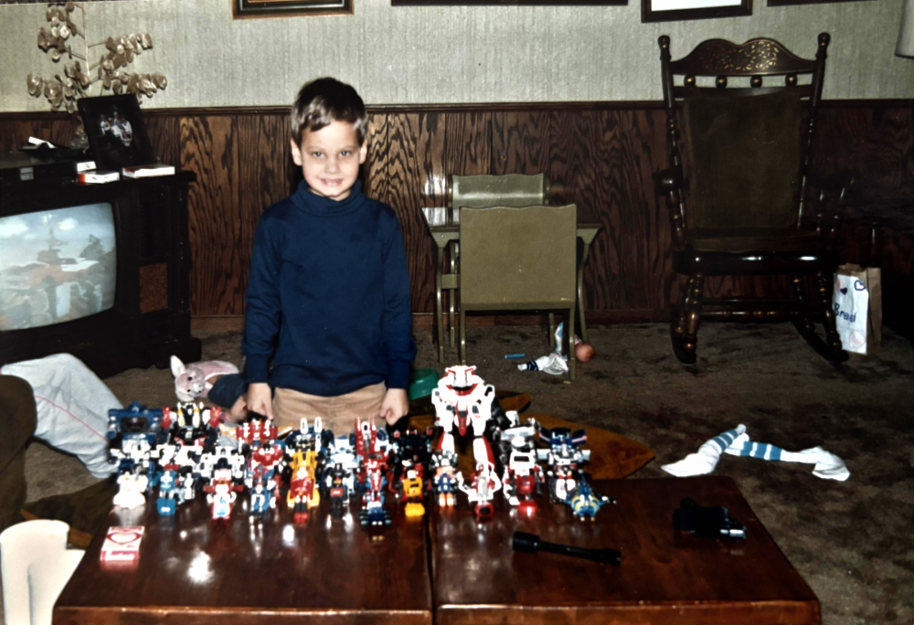 A young boy standing behind a coffee table covered with a large collection of robot action figures in a living room.