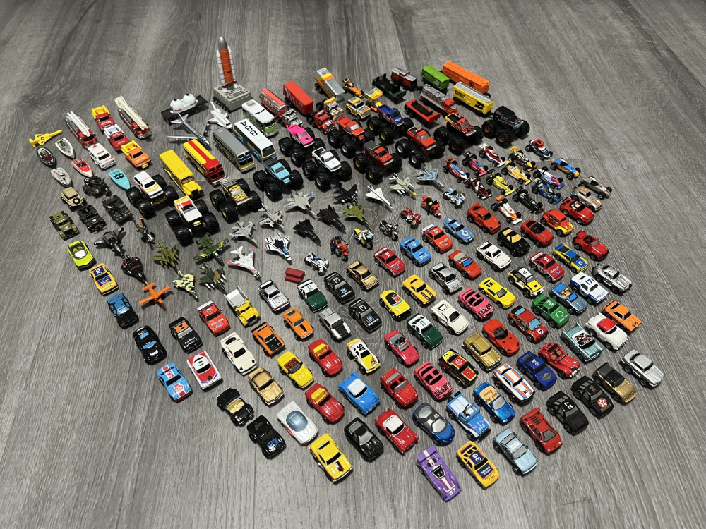 A large collection of toy cars, trucks, planes, and monster trucks arranged in rows on a gray wooden floor.