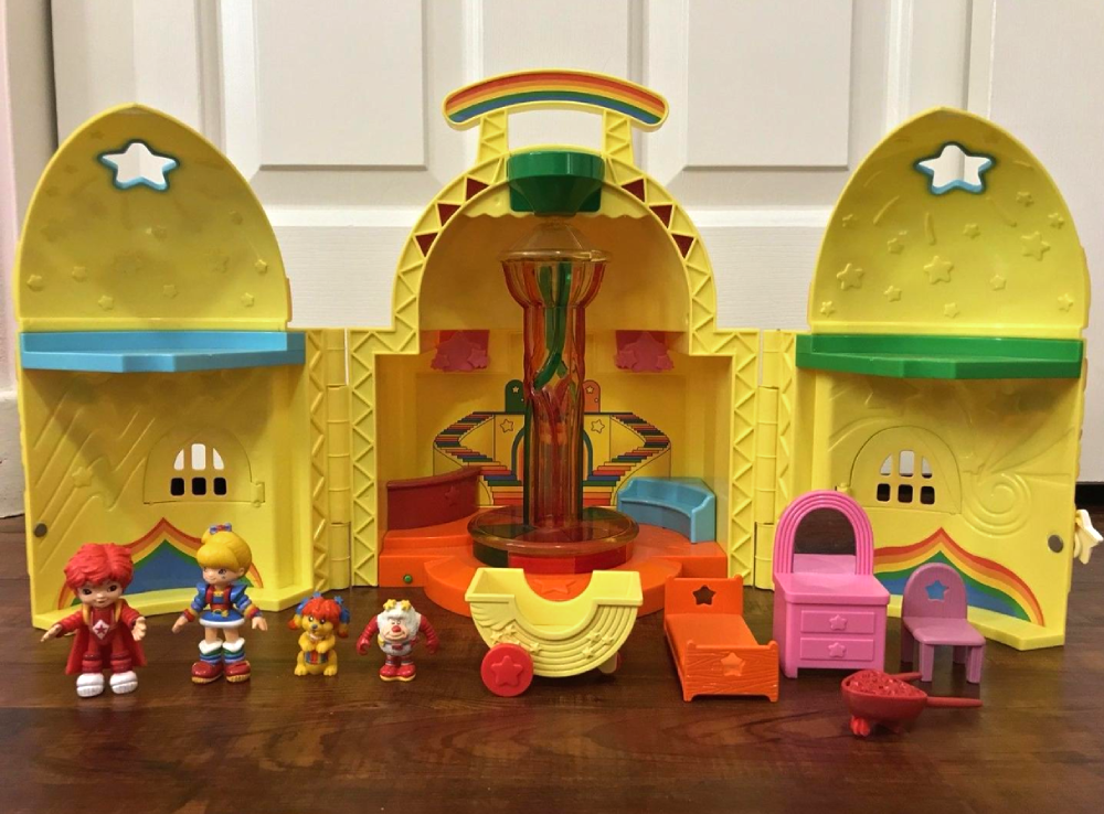 Rainbow Brite toy castle playset with several small character figures, furniture, and accessories arranged in front of it on a wood floor.