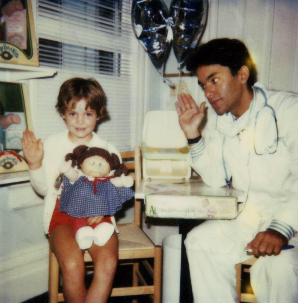 A young child holding a Cabbage Patch Kids doll sits next to a doctor in a lab coat, both waving.