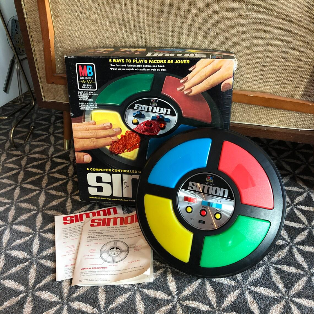 A circular Simon electronic memory game, its original box, and instruction manual.