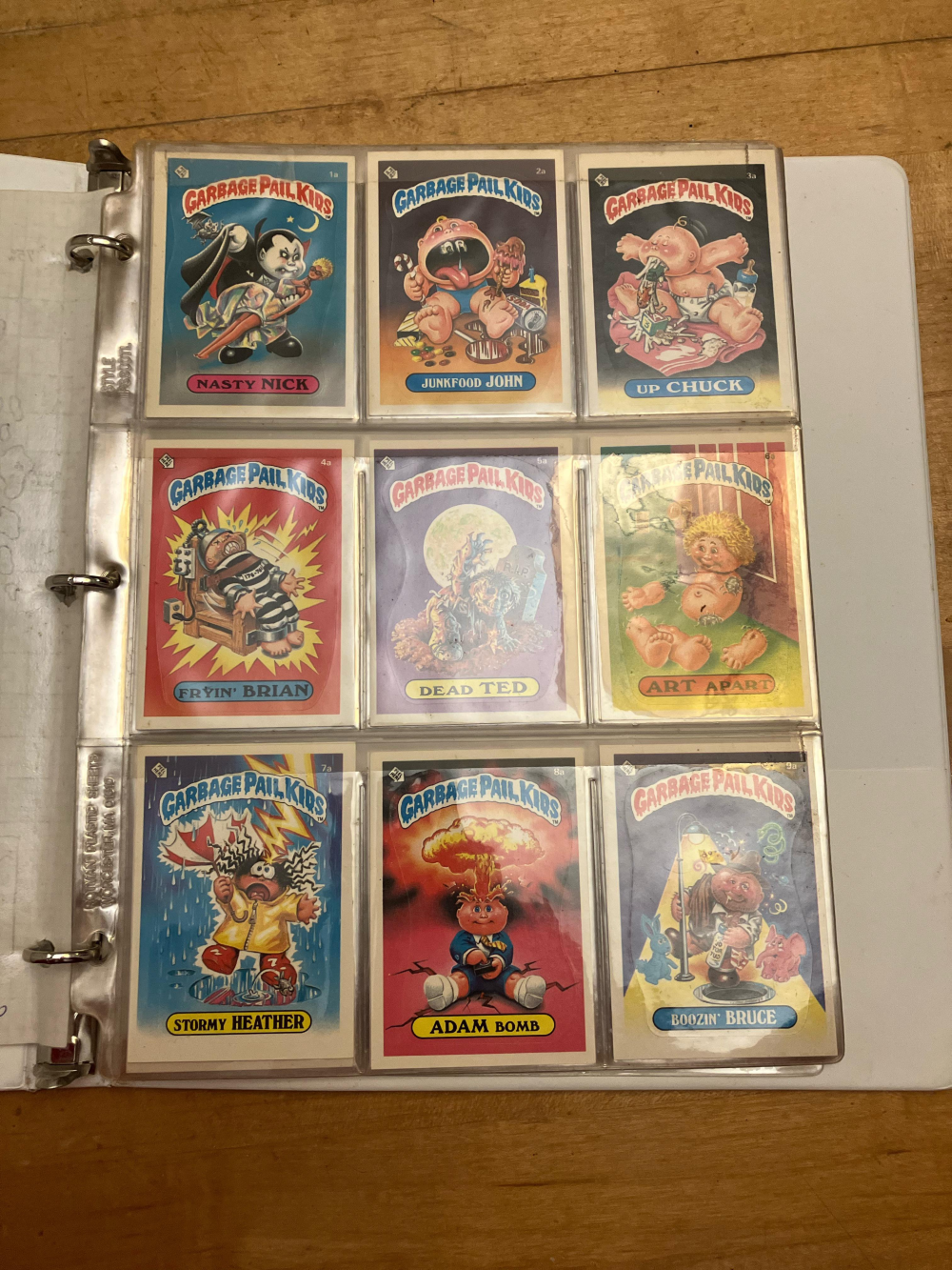 A plastic binder page containing nine vintage Garbage Pail Kids trading cards, including Adam Bomb, Nasty Nick, and Fryin’ Brian.
