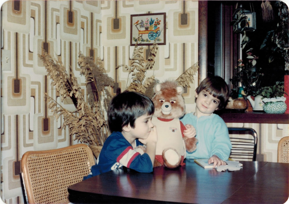 Two children sit at a table with a teddy bear between them.