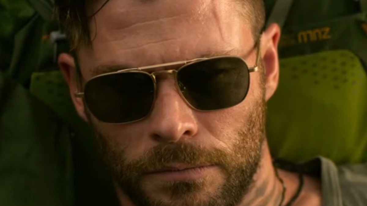 A man with a beard and sunglasses looks serious. He is wearing a green backpack and is outdoors.