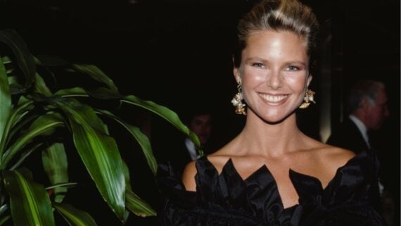 Christie Brinkley smiling while wearing a black off-the-shoulder dress and large statement earrings.