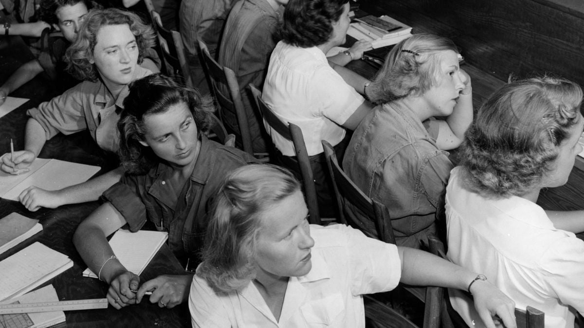 Black-and-white photo of several women seated at desks, writing in notebooks and turning to look toward the right side of the image, possibly listening to someone speak.