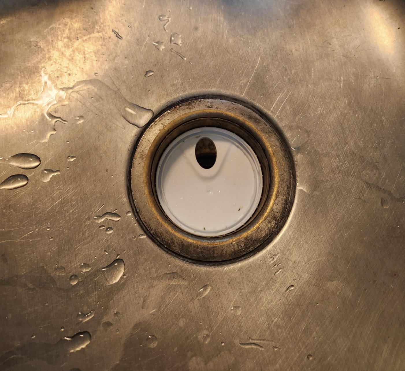 A close-up of a stainless steel sink with water droplets and a white drain cover at the center, surrounded by a slightly tarnished metal ring.