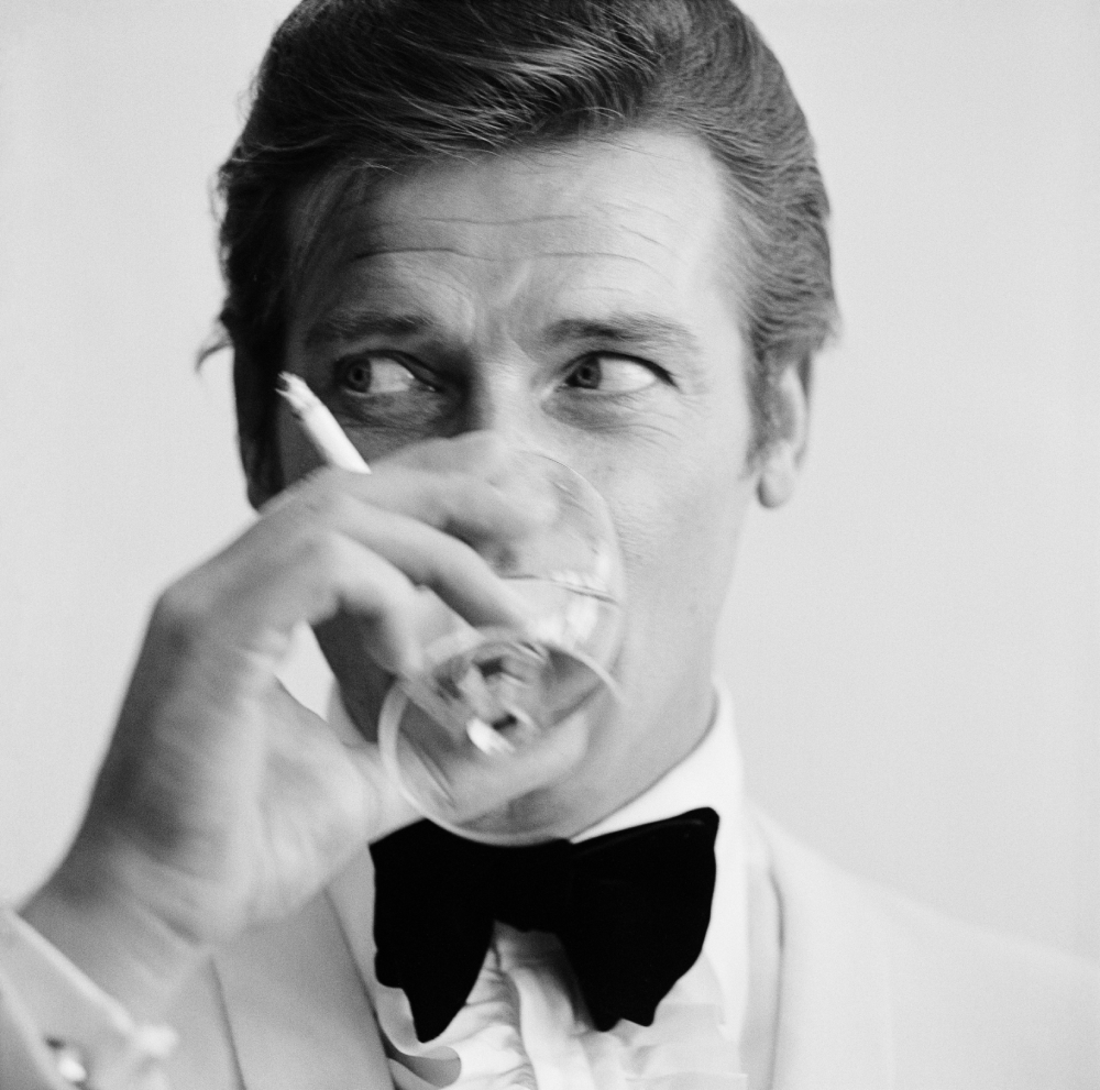 Roger Moore in a tuxedo and bow tie holding a glass and a cigarette, looking to the side.
