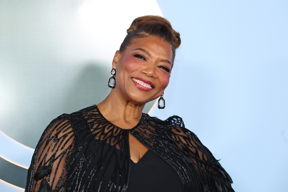 Queen Latifah smiling while wearing a black embellished cape and dangling earrings.