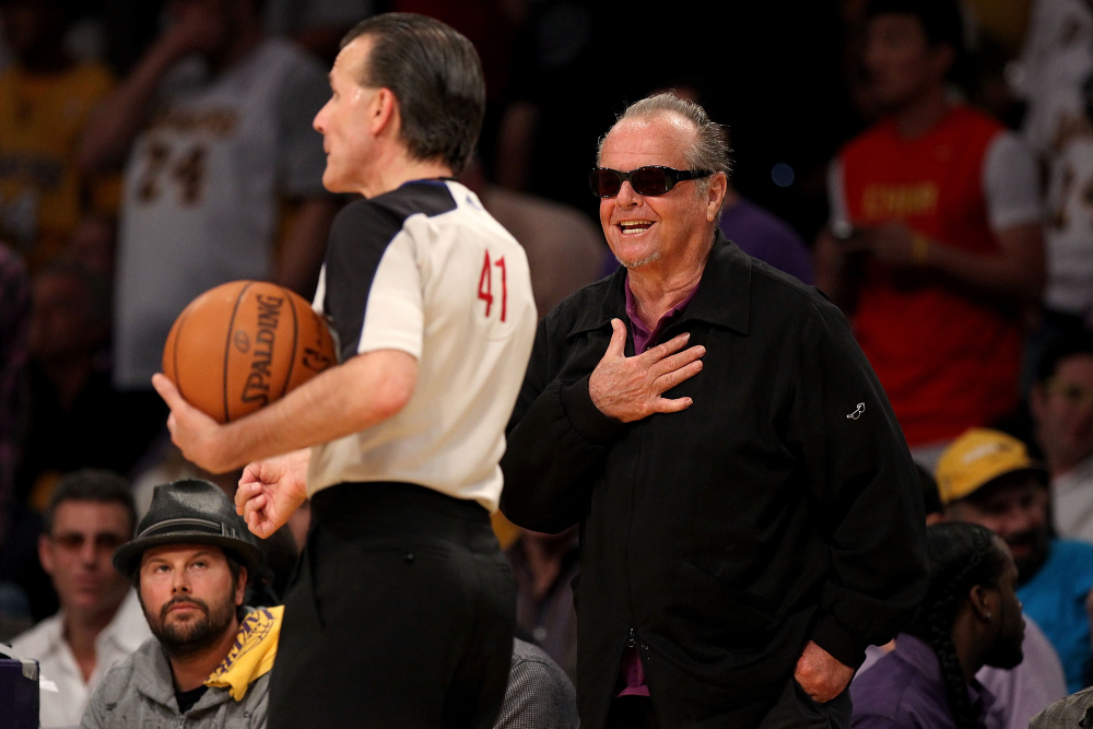 Jack Nicholson, wearing sunglasses and a black jacket, smiles with his hand over his heart while standing courtside near a basketball referee.