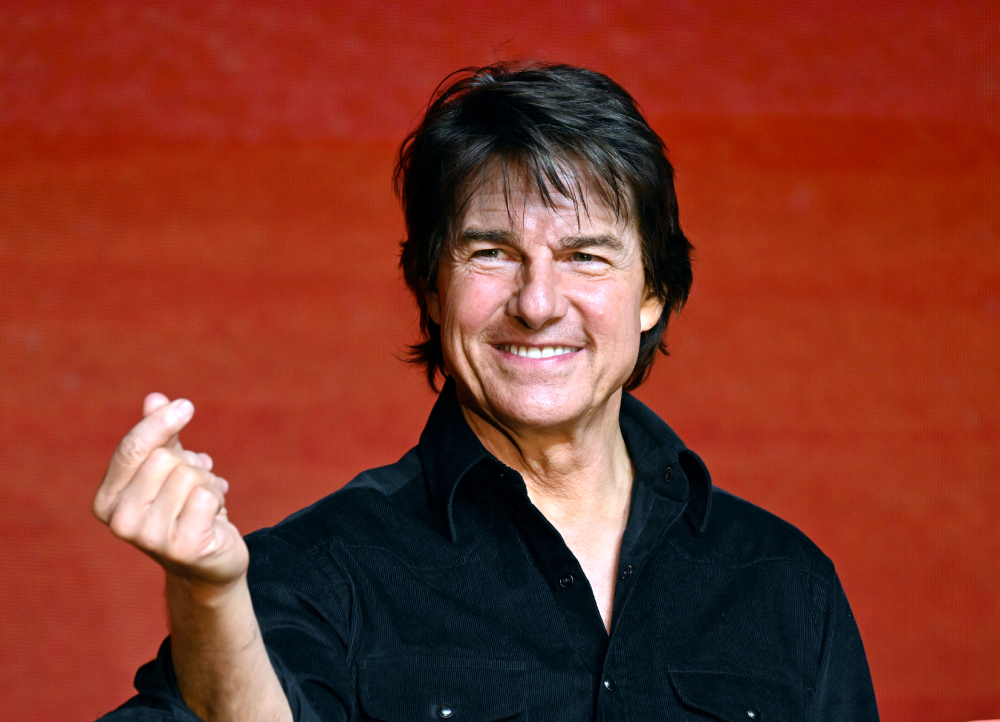 Tom Cruise smiling and making a finger heart gesture against a red background.