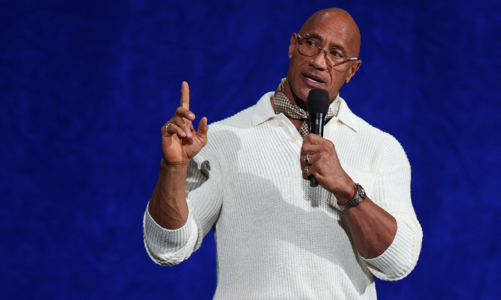 Dwayne Johnson wearing glasses and a white polo shirt speaks into a microphone while gesturing with one finger.