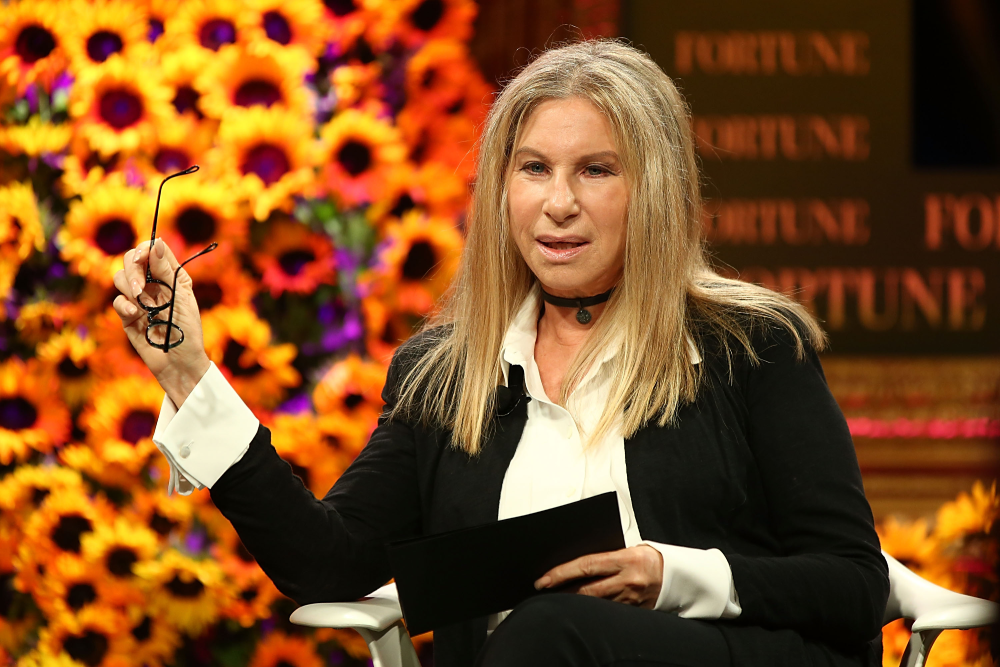 Barbra Streisand speaking at a Fortune event, holding a pair of glasses in front of a backdrop of sunflowers.