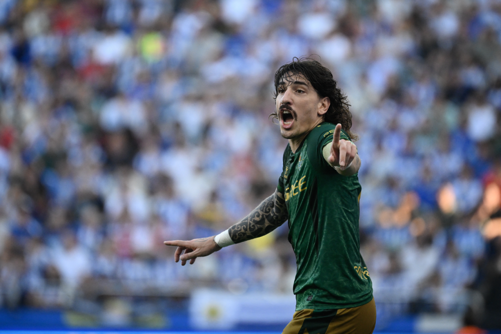 A soccer player in a green jersey gesturing with his arms while looking toward the side of the field in front of a blurred crowd.