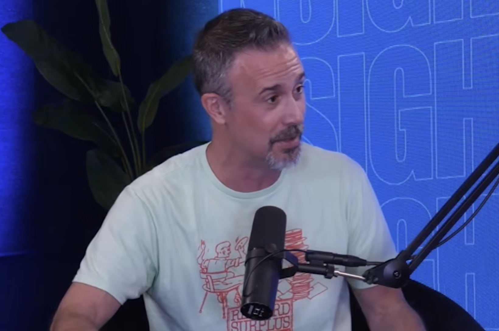 A man with a goatee wearing a light green t-shirt speaks into a microphone during a podcast recording.