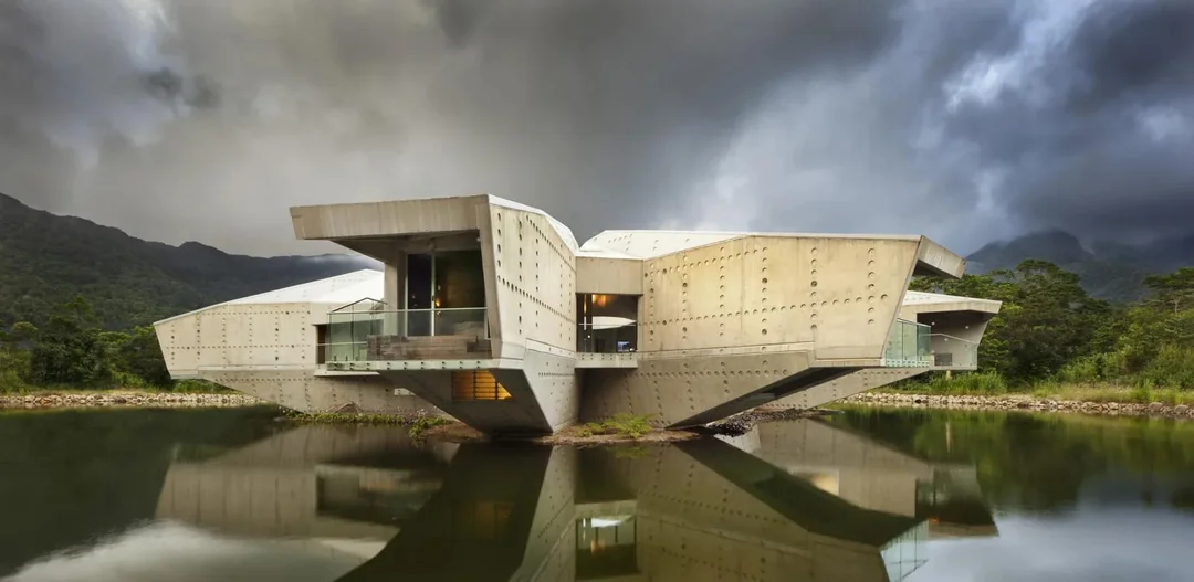 A modern concrete building with angular, cantilevered sections floating above a still pond in front of mountains.