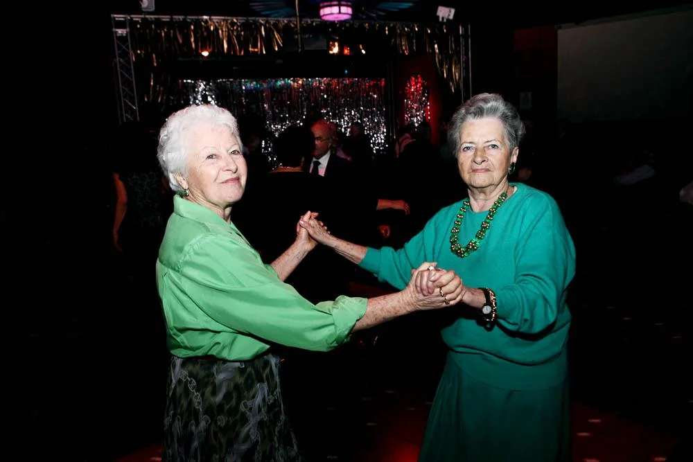 Two older women in green tops hold hands and dance together at a nightclub.