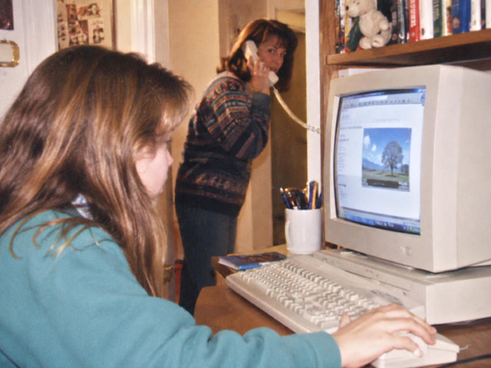 A person using a desktop computer while a woman in the background talks on a landline phone.