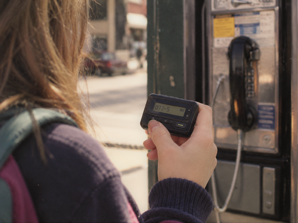 A person holding a pager with the number 077-5 on its screen while standing next to a public payphone.