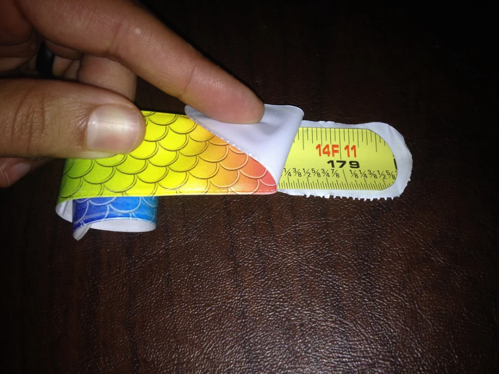 A hand peeling back a colorful, fish-scale patterned sticker to reveal a yellow measuring tape underneath marked “14F 11” and “179”.
