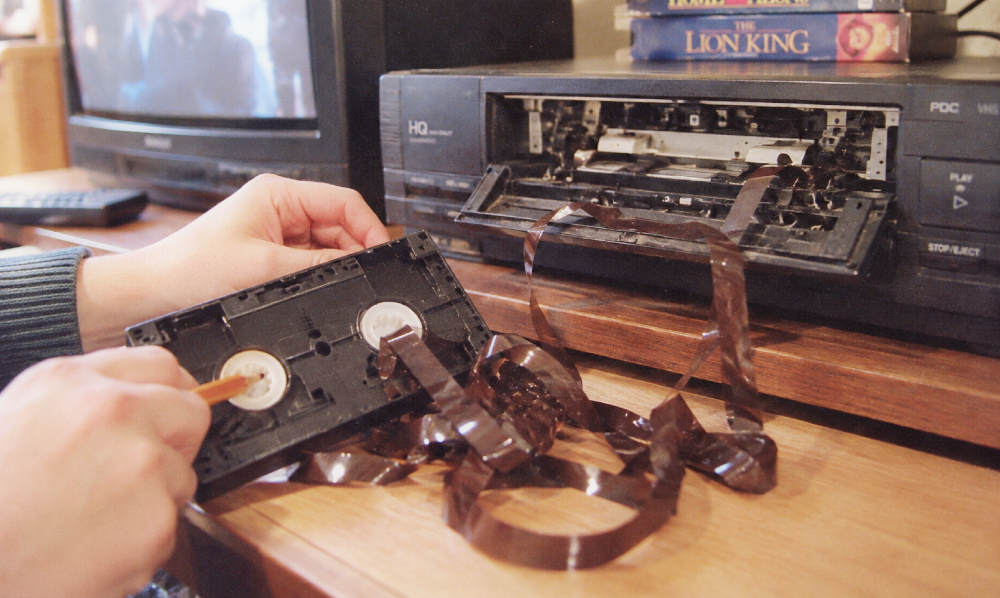 Hands using a pencil to manually wind magnetic tape back into a VHS cassette next to an open VCR.