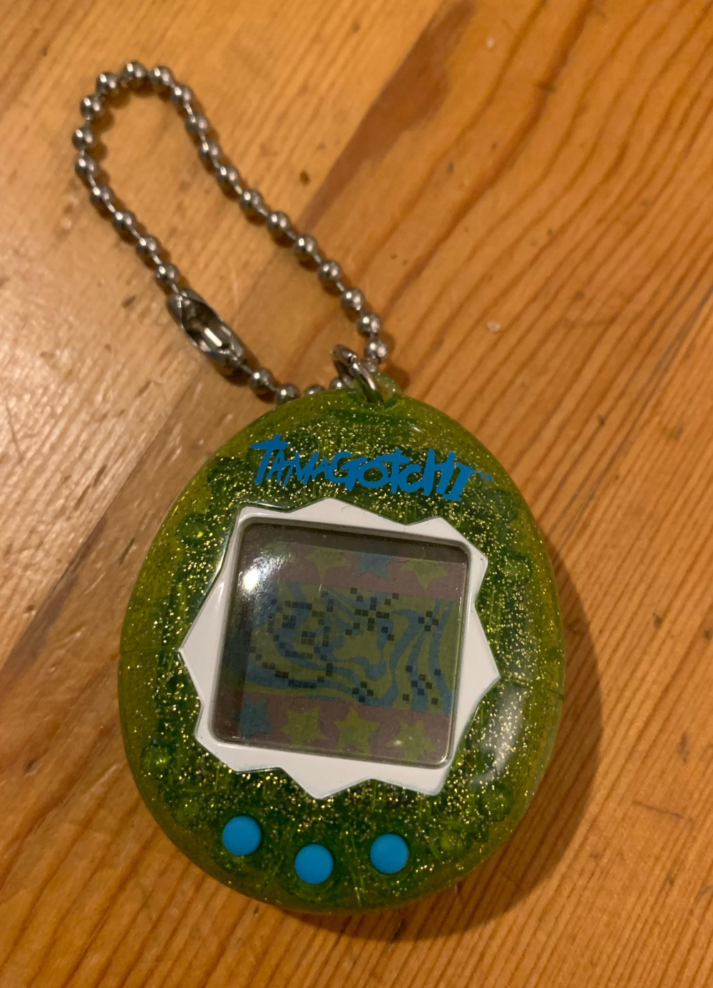 Green glittery Tamagotchi keychain on a wooden surface.