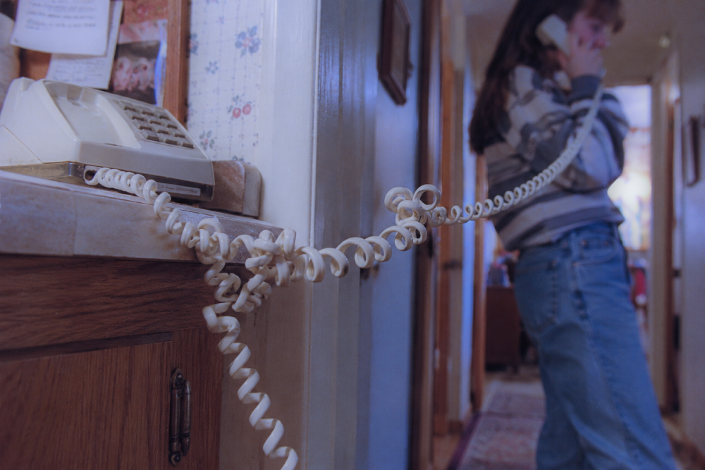 A person stands in a hallway talking on a wall-mounted landline phone with a long, tangled coiled cord.