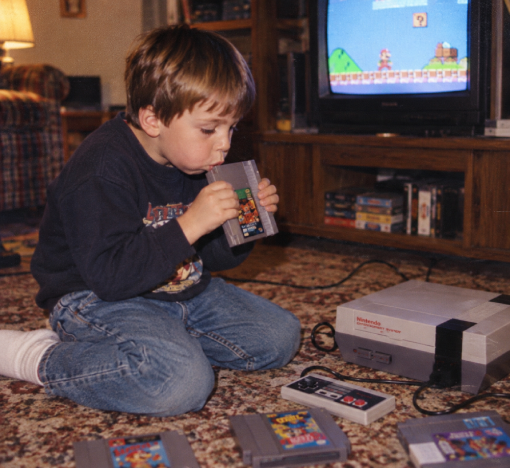 A young boy kneeling on a carpet blows into a Nintendo game cartridge in front of a television displaying Super Mario Bros.