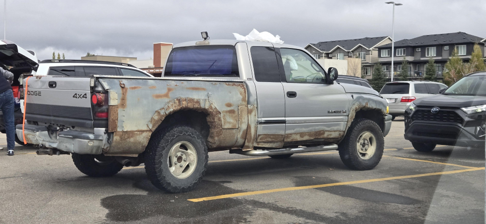 A silver Dodge pickup truck with heavy rust along the rear wheel well parked in a parking lot.