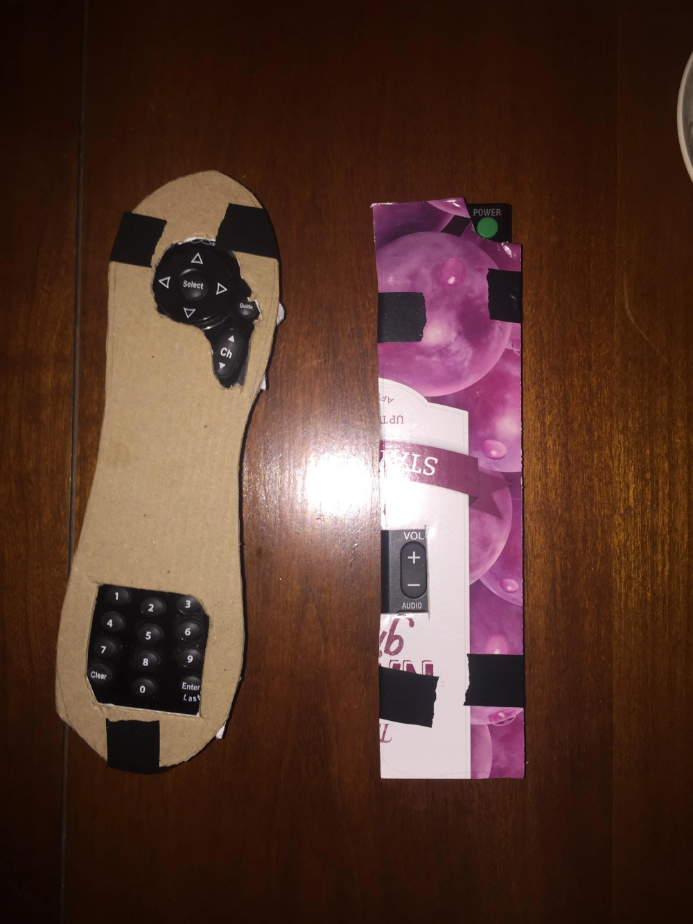 Two homemade TV remotes constructed from cardboard, paper, and salvaged remote control buttons, resting on a wooden surface.