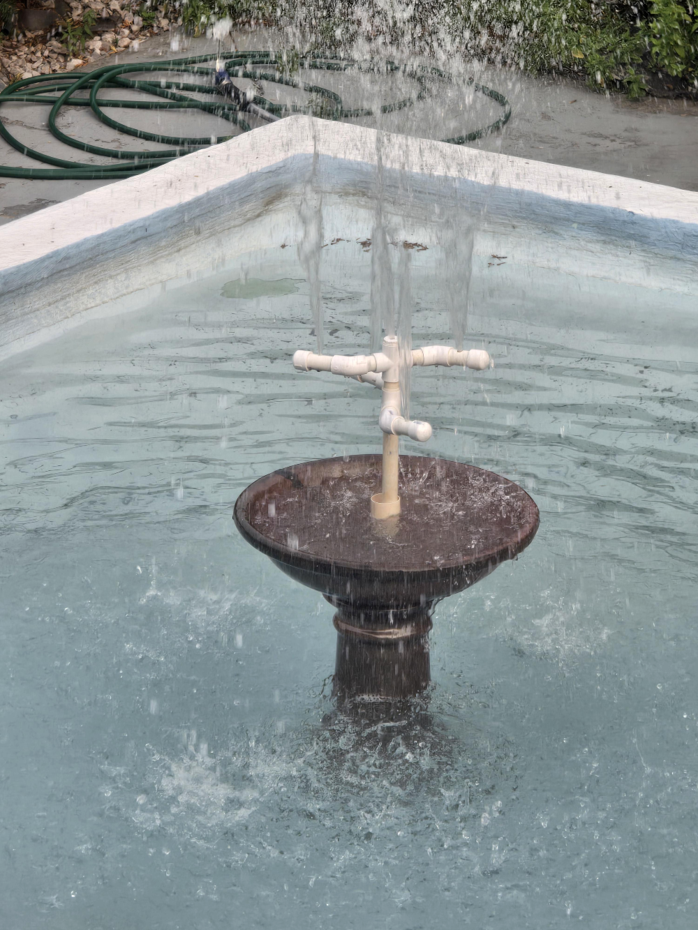 A small fountain with a PVC pipe sprinkler head spraying water into a tiered basin.