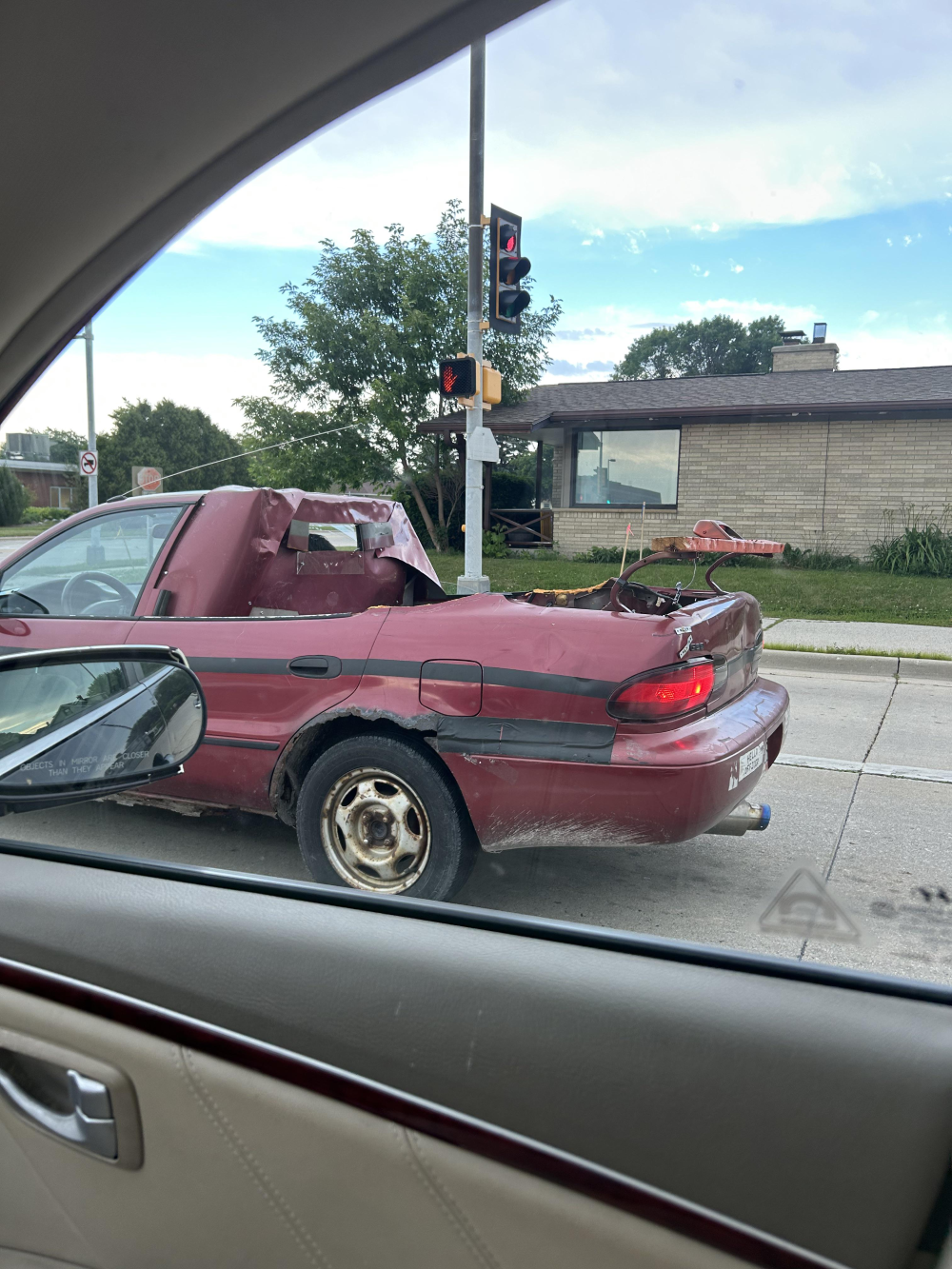A heavily damaged red sedan with a crushed roof and removed trunk lid driving on a city street.