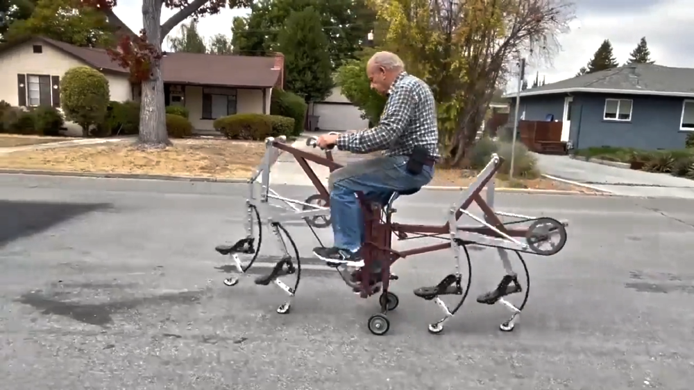 A man rides a custom-built mechanical bicycle that uses walking legs instead of wheels to move along a residential street.
