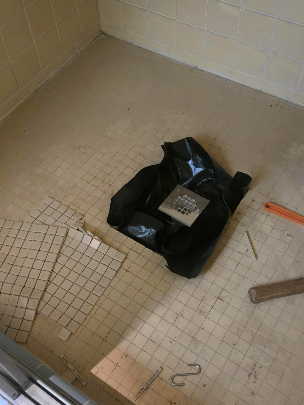 A shower floor under construction with a central drain, black waterproof membrane, and loose sheets of square mosaic tiles.