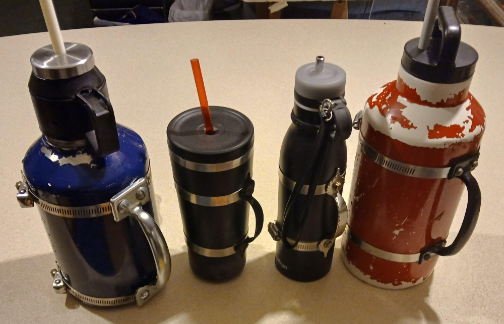 Four insulated beverage containers of varying sizes and colors sitting on a table, each featuring a custom-attached metal handle secured with hose clamps.